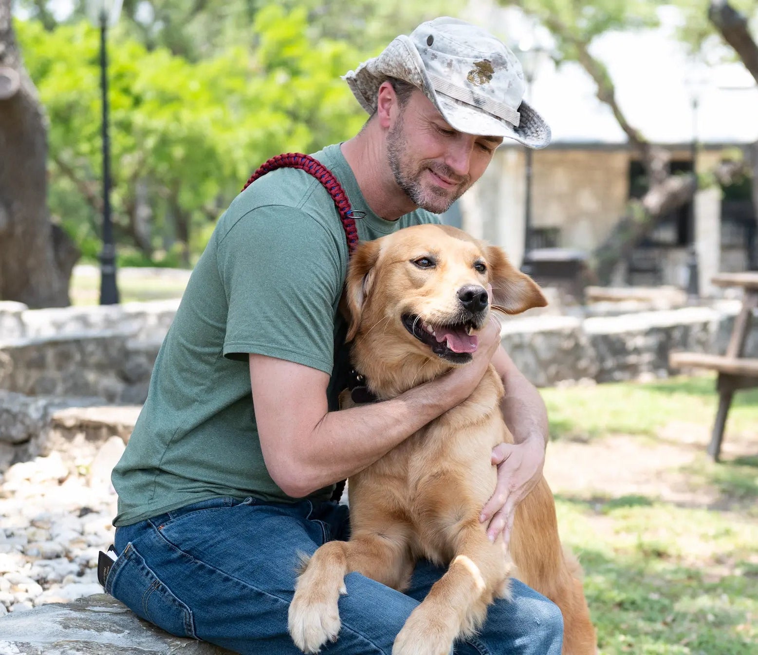 Man sitting outdoors with a dog on his lap, surrounded by greenery