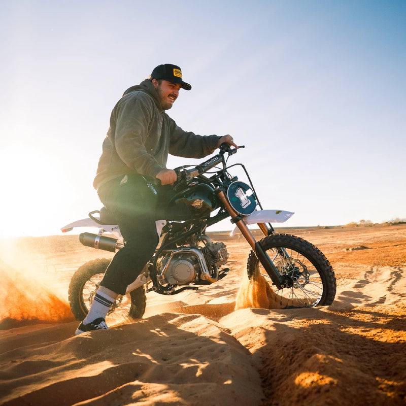 A rider on the black pit bike on a sandy trail with the sun low behind him