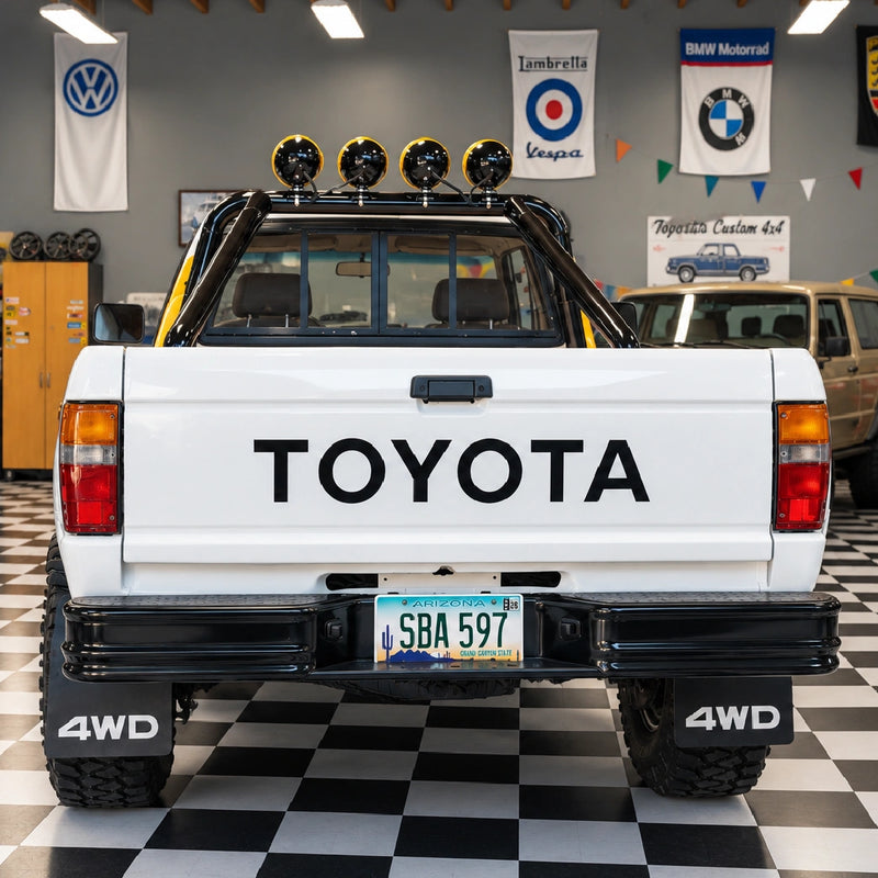 The back of the white Toyota with black tailgate lettering, a black rear bumper, 4WD mud flaps and four lights on the roll bar