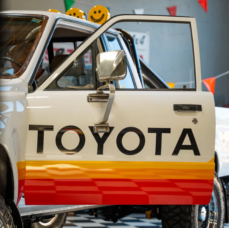 The open door of the white Toyota showing black Toyota lettering, striped paint and a chrome side mirror